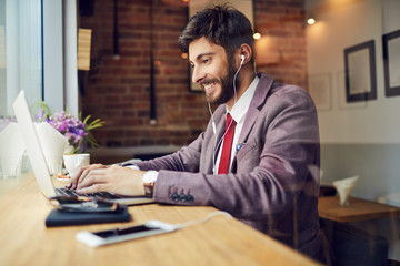 Cheerful young businessman working on laptop and listening to music while sitting at table in a cafe