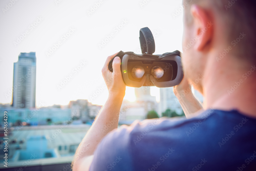 Wall mural VR Goggles. Man ready to wear virtual reality goggles. The vr headset design is generic and no logos. Backward light, selective focus, close up. Sunset city landscape.