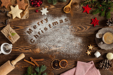 Wording Merry Christmas written on dark wooden background, copy space. Cookies, candle, cinnamon, cup of coffee, marshmallows, fir-tree, dried oranges. Top view.