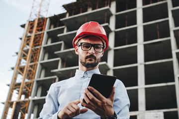 Young business man builder in red hard hat with tablet in hand. Portrait engineer or architect with...