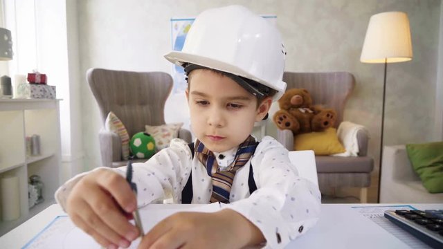 Small Kid Boy In White Helmet Play In Constructor, Imitating Grown Up Working And Busy Man, Using Pencils And Pair Of Compasses, Drawing Plan On Paper Sitting At Table And Being Very Concentrated