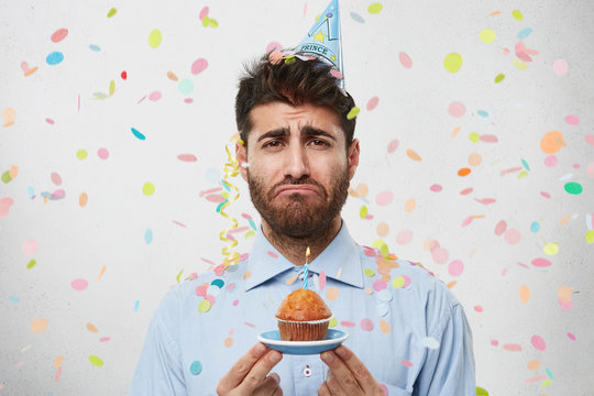 Picture of upset unshaven man in blue shirt feeling unhappy about his 30-year anniversary, screwing mouth and looking at camera with mournful expression, holding cupcake with one candle on it