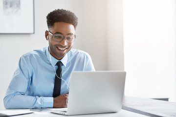 Indoor office shot of cheerful successful young African American manager with stubble sitting in front of open laptop wearing earphones while having video conference call with business partners