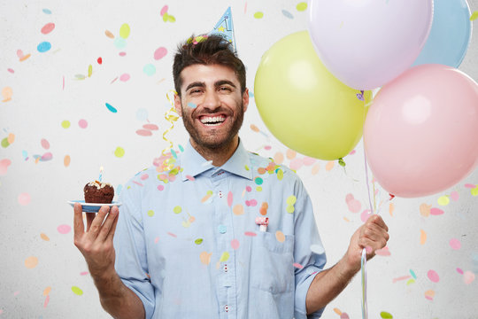 People, joy, fun and happiness concept. Relaxed happy birthday guy looking cheerful, smiling happily, posing for picture, holding colorful helium balloons and cupcake with confetti falling down