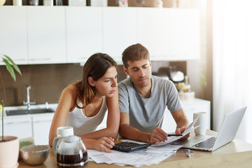 European couple sitting together indoors over home interior, calculating finances at home, looking...