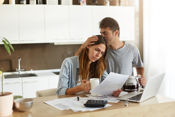 Frustrated woman studying sheet of paper in kitchen, drinking coffee and trying to find way to pay...