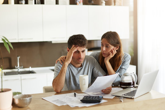 Depressed young Caucasian couple calculating family budget in kitchen, trying to find a way to make both ends meet, having stressed and frustrated looks: caring wife embracing her unhappy husband