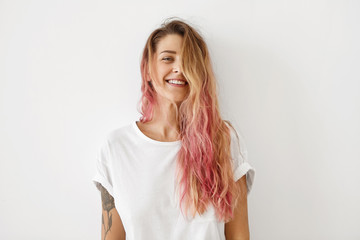 Fototapeta Horizontal portrait of pleasant-looking Caucasian female with long hair, pink on tips, having tattooes on arms, wearing white casual T-shirt, covering her face with hair, looking happily in camera obraz