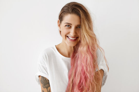 Pleasant-looking cheerful young female with blue eyes, long luxuriant hair, tattooed arms, wearing oversize T-shirt, looking directly into camera with broad smile, demonstrating her white teeth