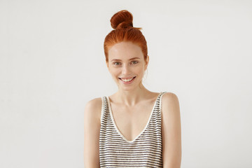 Happy freckled woman with red hair knot, wearing loose striped shirt, feeling relaxation while resting at home, having good mood, smiling pleasantly, showing her perfect white teeth. Youth, lifestyle