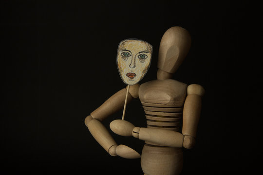 Wooden doll on hinges holds a mask in hands and covers her face on a black background