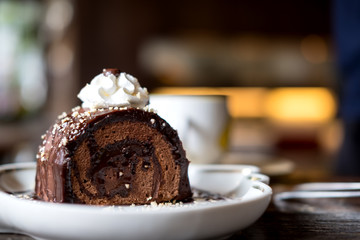 A piece of chocolate roll cake and whipped cream on the top in white ceramics plate and coffee cup on wooden table with blur background in cafe