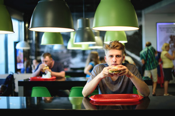 Young man sits in a fast-food restaurant with a burger in his hands. Delicious food for lunch.