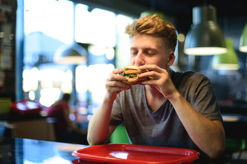 Student eats a delicious burger in a fast-food restaurant and receives pleasure from it.