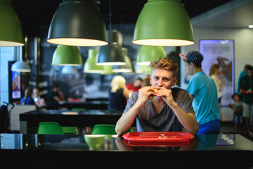 Student greedily eats a great beautiful burger sitting in a fast food restaurant.