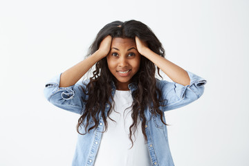 Naklejka premium Beautiful Afro-american woman in casual clothes with long wavy hair looking nervously holding her hands on head feeling distrust or fear while posing against white background.
