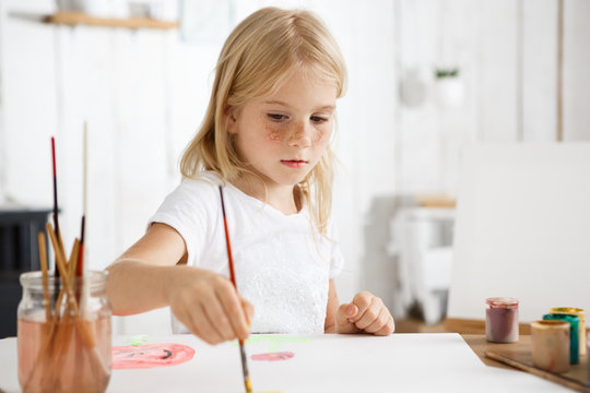 Close up portrait of little white-skinned girl with blonde hair and freckles focused on painting picture for her parents. Blond baby sitting at the desk with brush in her hand.