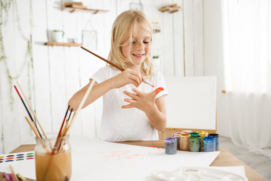 Beautiful, creative and busy little blonde girl in white t-shirt drawing on her palm with a brush. Eight-year-old freckled female kid standing behind desk at the art room filled with sunlight. Kids