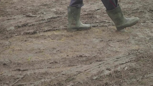 Man's legs in rubber boots walking on mud from left to right.