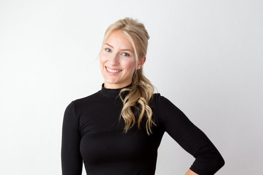 Junge Frau mit jugendlichem Aussehen im schwarzen engen Pullover vor einer wei&szlig;en Wand mit sch&ouml;nen blonden langen gesunden Haaren