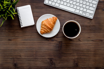 Light lunch in office. Coffee and croissant near keyboard and notebook on dark wooden background top view copyspace