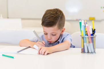 child preschooler boy drawing on booklet ,while sitting on the desk in the classroom