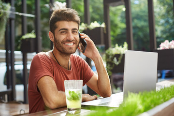 Shot of male smiling to the camera good-looking freelancer working online via laptop at the cafe, discussing work on the cell phone. Bearded guy in red t-shirt sitting in front of notebook sitting at