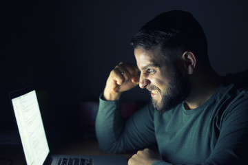 Obraz premium Happy young man celebrating success with raised fist in front of the computer. Working late at night. 