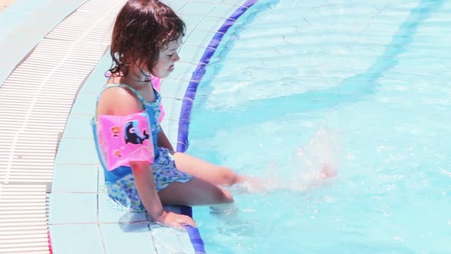 Young girl in swimsuit sitting on pool border and playing in water