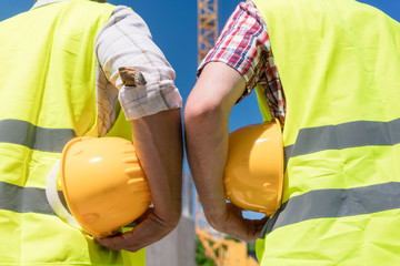 Close-up rear view of the hands of two workers wearing reflective safety vests while holding yellow...