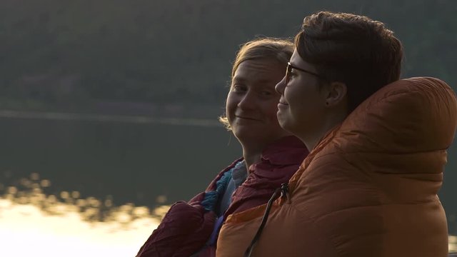 Cute Couple Relax In Their Sleeping Bags And Enjoy Sunset At River, They Cuddle