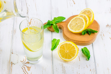 Fototapeta premium In a glass beaker, a cold lemonade is poured from a jug on a white wooden background surrounded by lemons.