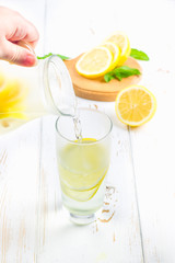 In a glass beaker, a cold lemonade is poured from a jug on a white wooden background surrounded by lemons.