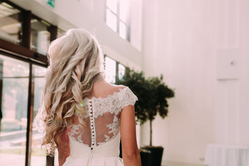 Close-up image of back of elegant blonde with long curly hair and luxury wedding dress. Bride's morning