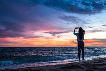 Young woman traveller making heart shape on the beach at beautiful sunset