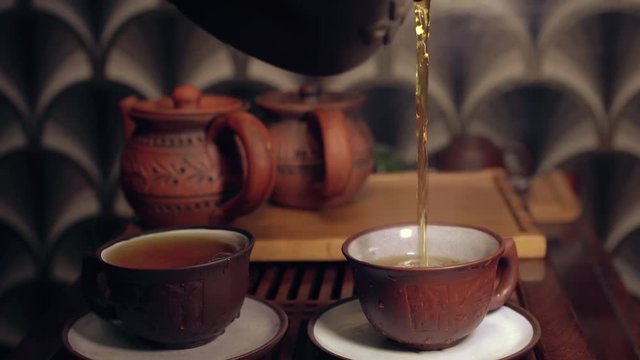 The action of traditional Chinese tea ceremony. Pouring tea from clay tea pot in a cup. Two tea pots in soft focus on the background.