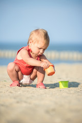 Baby playing on the sandy beach near the sea. Cute little girl in red dress with sand on tropical beach. Ocean coast.