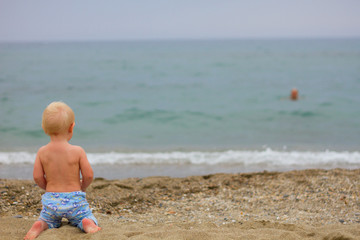 Blond baby sitting on the beach/Blond baby wearing a blue pants sitting on the beach, waitting for someone from the sea on a cloudy day