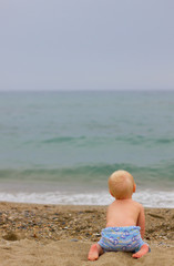 Fototapeta na wymiar Blond baby sitting on the beach/Blond baby wearing a blue pants sitting on the beach, waitting for someone from the sea on a cloudy day