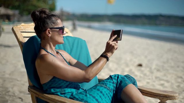 Beautiful woman sitting on the sandy beach and doing photos on smartphone, steadycam shot
