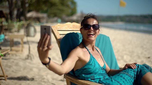 Beautiful woman sitting on the sandy beach and doing selfies on smartphone, steadycam shot
