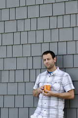 Man leaning against a gray wall holding an orange coffee mug outside. 