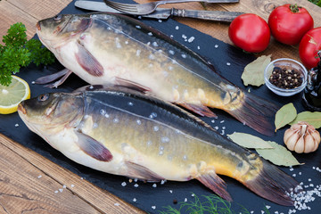 Fresh raw river mirror common carp fish on black stone cutting board on wooden table. Food ingredients for preparing. Cooking and kitchen concept background. Healthy seafood. Lemon, herbs and spices.
