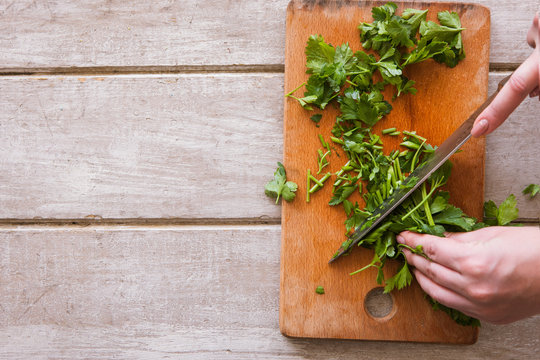 Woman cuts batch of parsley on wooden desk with steel knife. Herbs as healthy and organic food, top view picture, free space nearby