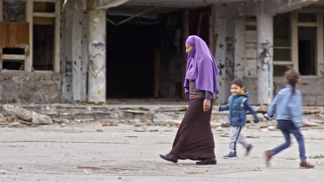 Tracking of middle-eastern woman in burqa carrying groceries and walking through street with abandoned building while her children running and playing; soldiers with firearms patrolling in background