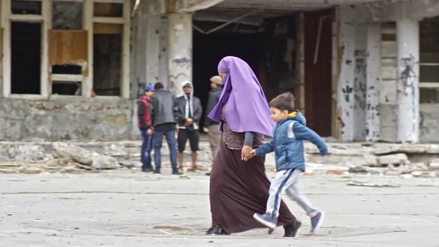 Tracking of Muslim woman in purple niqab holding hands of little boy and girl and walking along street with abandoned building; soldiers with firearms patrolling in background