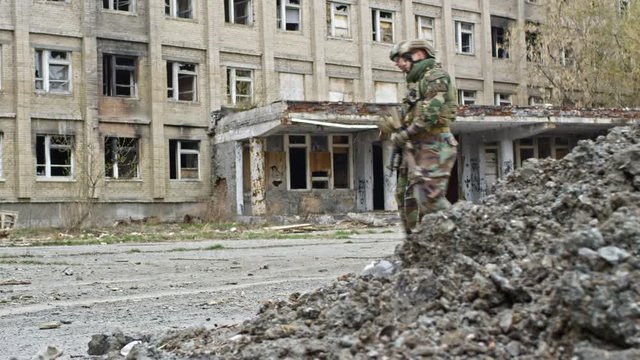 PAN of soldiers in military camouflage carrying firearms and patrolling dirty street, then male refugees and female Muslim woman with children walking towards abandoned building with broken windows