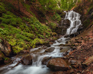 Fototapeta premium Majestic waterfall amid the dense lush green, with flowing stream and boulders in the foreground. Blurred effect on water. Carpathian, Ukraine
