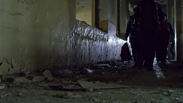 PAN with low angle of group of male refugees and Muslim woman in niqab holding hand of little girl walking along dirty floor in dark abandoned building; unrecognizable soldiers in background