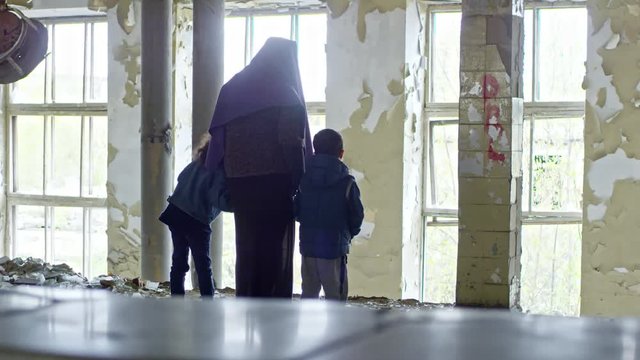 PAN with rear view of Muslim woman in purple niqab standing before broken window in abandoned building and holding hands of little children: she pointing and showing something to them